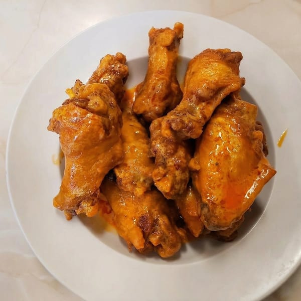 A white plate piled high with golden-brown chicken wings coated in a vibrant orange buffalo sauce.